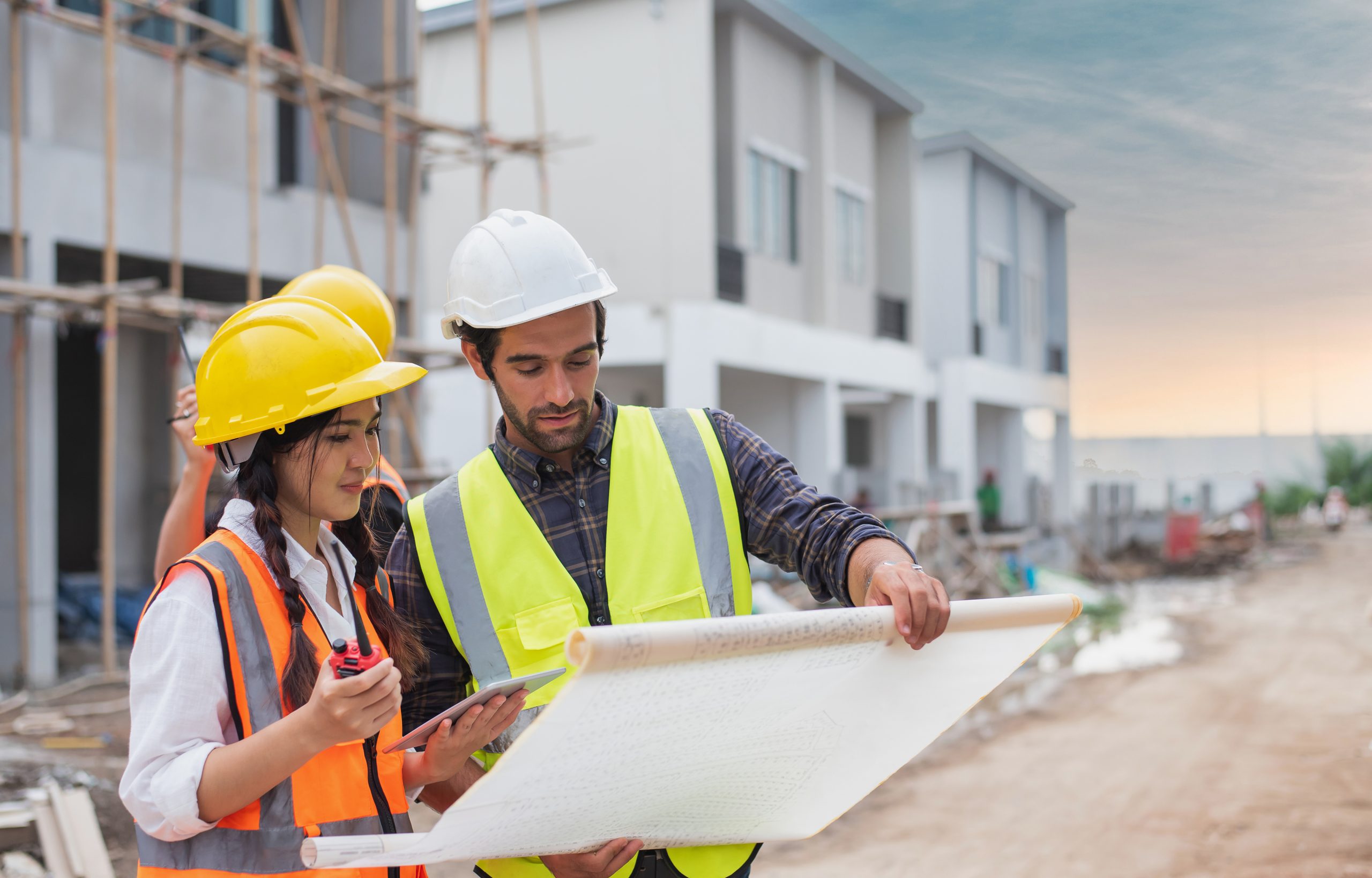 caucasian males and asian female builders, architector and engineers with draft plan of building and laptop computer talking on constructing site